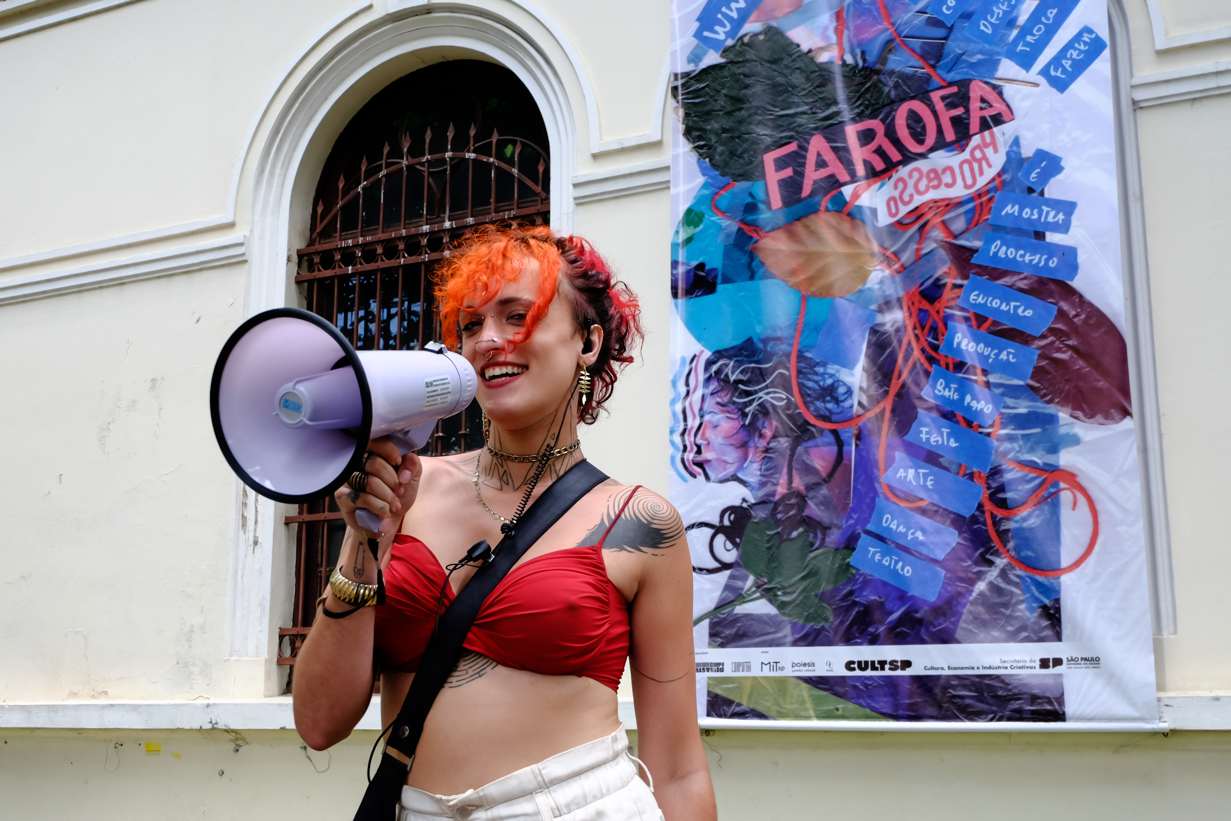 Gabs Ambròzia em foto de Camila Rios, em frente à Oficina Cultural Oswald de Andrade, com o banner do Farofa do Processo ao fundo