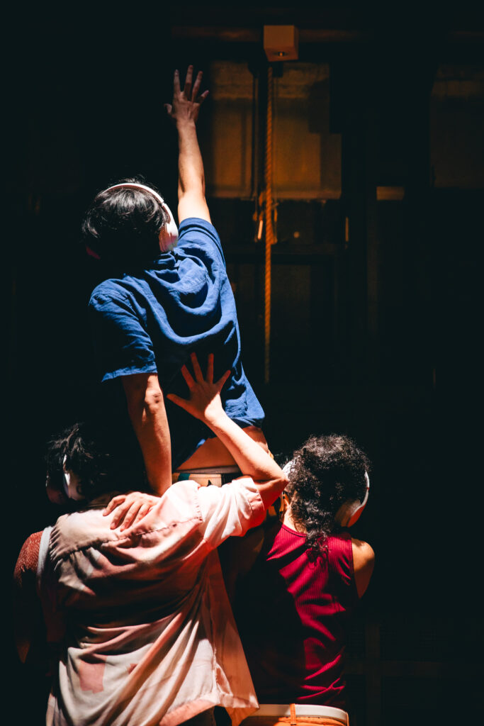 Foto de cena do espetáculo "Coro dos que dão as costas". Três pessoas de costas, utilizando fones de ouvido. Duas seguram o terceiro sobre seus ombros, e este, no alto, estende seu braço direito para cima, como que tentando alcançar algo.